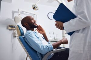 Man in blue shirt in dentist's chair holding one hand to his face smiling