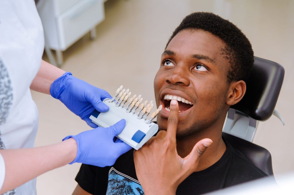 Man in dental chair pointing to his teeth as dentist holds shade guide up